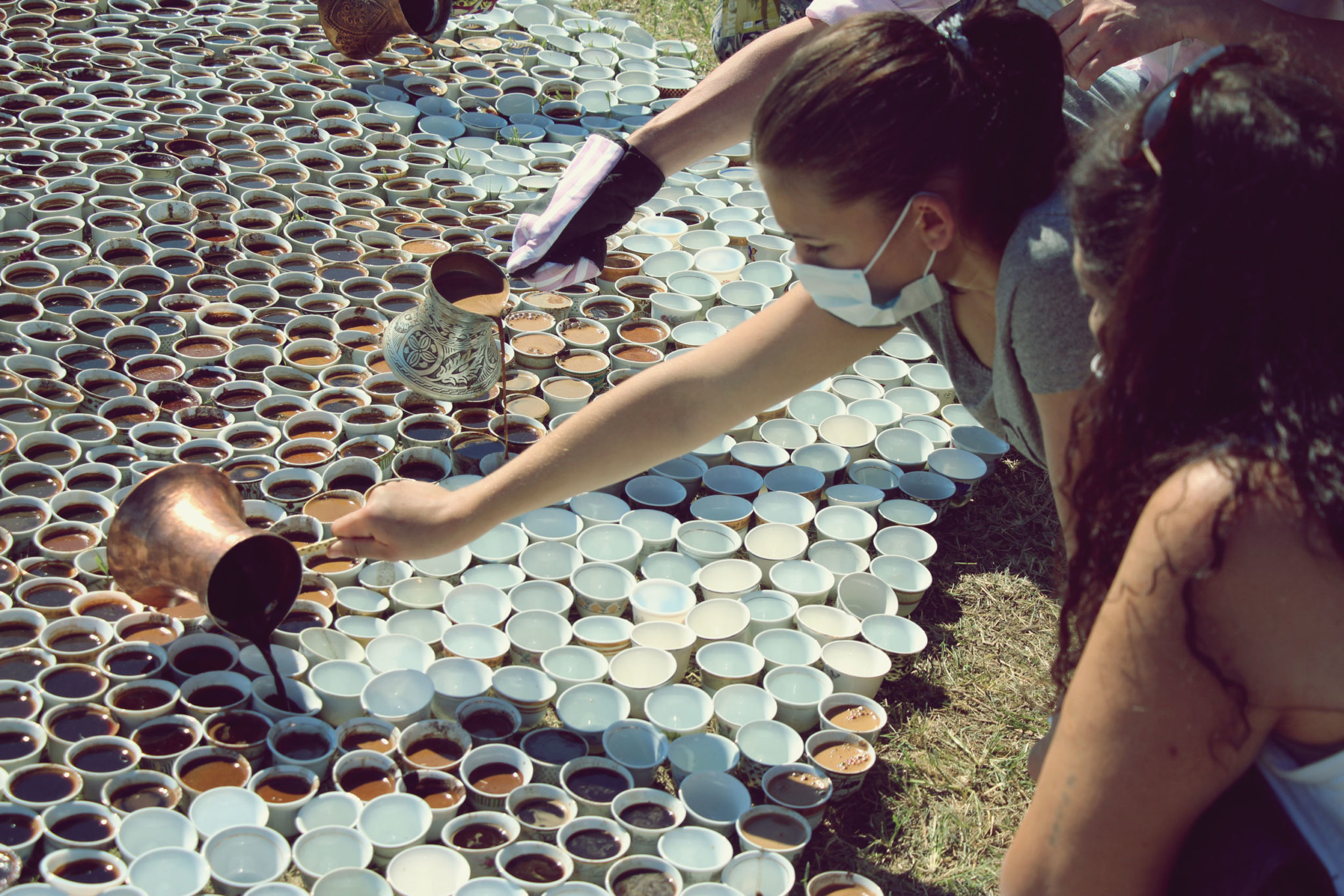 Various people sit on the ground in front of a large number of white cups, more than can easily be counted, that are closely packed together. Roughly half of the cups are filled with a brown liquid. Three people are pouring more coffee from metal containers into cups.